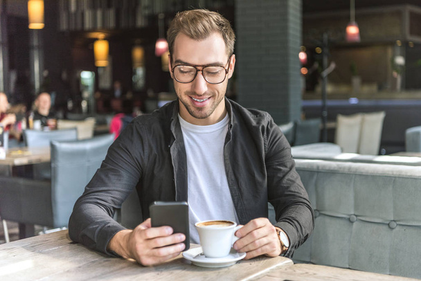 smiling young man with cup of coffee using smartphone in cafe - Фото, зображення