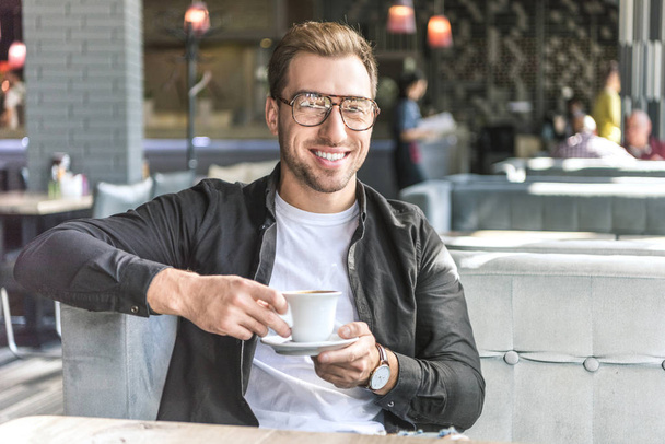 happy young man with cup of coffee sitting in cafe and looking at camera - Фото, зображення