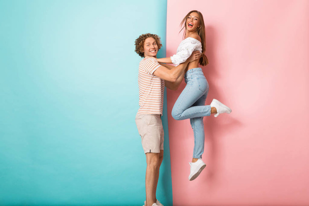 Full length portrait of a happy young couple standing over two colored background, man lifting up his girlfriend - Valokuva, kuva
