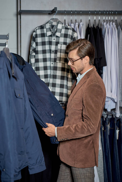stylish man in eyeglasses looking at jacket while shopping in boutique     - Photo, Image