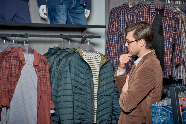 joven guapo con la mano en la barbilla mirando ropa elegante en boutique
 - Foto, Imagen