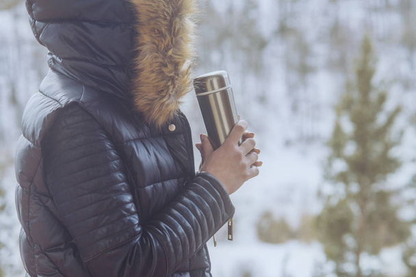 A girl in a winter jacket with a thermos of coffee or tea in her hand on the balcony in winter - Fotoğraf, Görsel