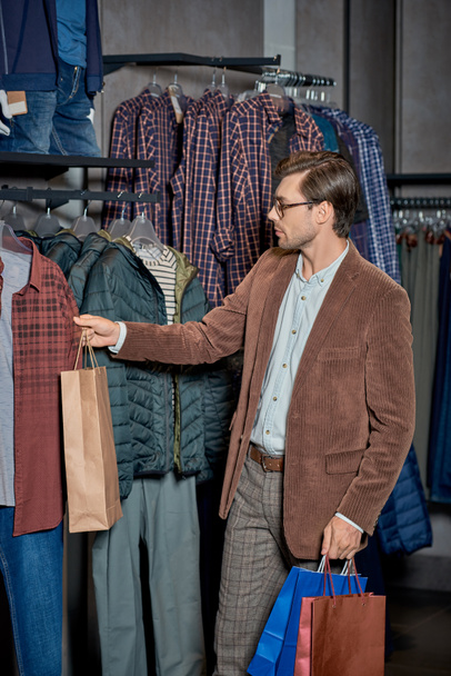 schöner Mann mit Einkaufstaschen und Kleiderauswahl im Geschäft - Foto, Bild