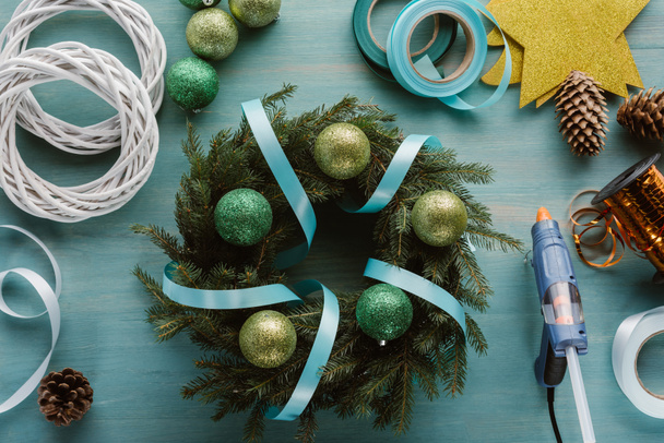 bovenaanzicht van gearrangeerde lijmpistool, chrismtas speelgoed en decoraties voor handgemaakte kerst krans op blauwe houten tafelblad - Foto, afbeelding