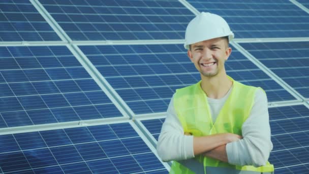 Portrait of a worker in overalls and a helmet on the background of solar panels. Smiles, looks into the camera - Felvétel, videó