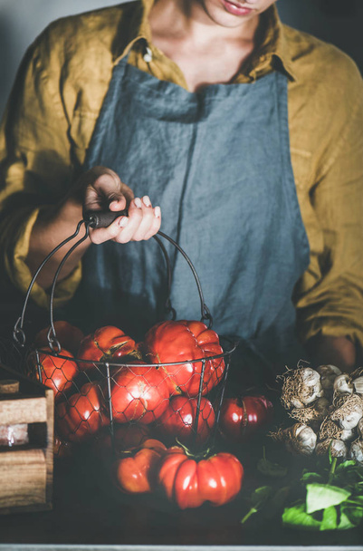 Woman in linen apron holding basket with ripe heirloom tomatoes for cooking tomato sauce, canned tomatoes or pasta with basil and garlic at kitchen counter. Healthy cooking, slow food or comfort food - Foto, Imagen