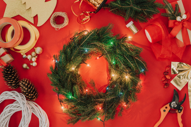 flache Liege mit Tannenzweigen, Weihnachtsbeleuchtung und Bändern für handgefertigten Adventskranz auf rotem Hintergrund - Foto, Bild