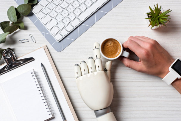 imagen recortada de hombre de negocios con prótesis mano y smartwatch sentado en la mesa con taza de café y teclado de ordenador en la oficina
  - Foto, Imagen