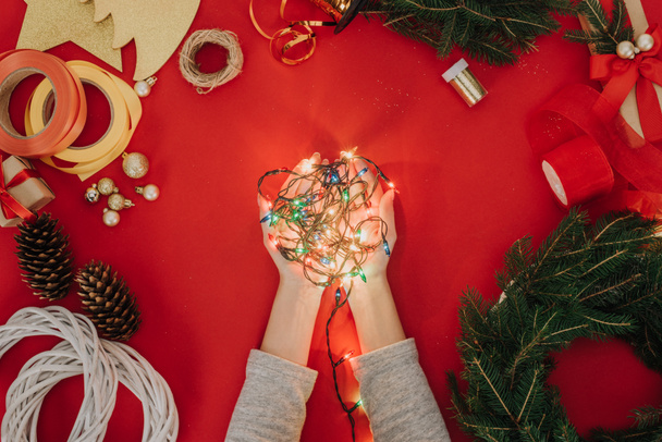 cropped shot of woman holding christmas lights in hands with pine tree branches and decorations for handmade wreath on red backdrop - 写真・画像