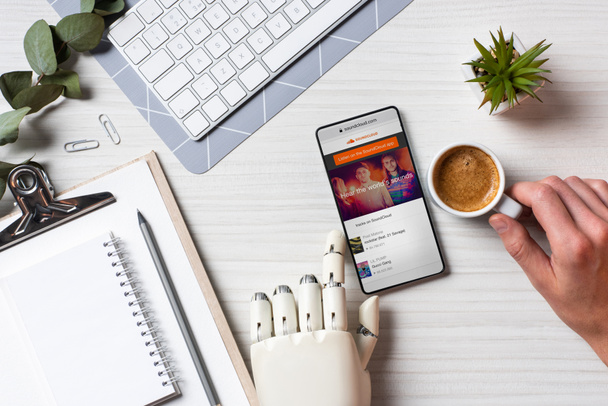 cropped image of businessman with prosthesis arm using smartphone with soundcloud on screen at table with coffee cup in office  - Фото, изображение