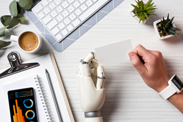 обрізане зображення бізнесмена з рукою кіборга і смарт-годинником, що тримає порожню візитну картку за столом з смартфоном з інфографікою на екрані в офісі
  - Фото, зображення