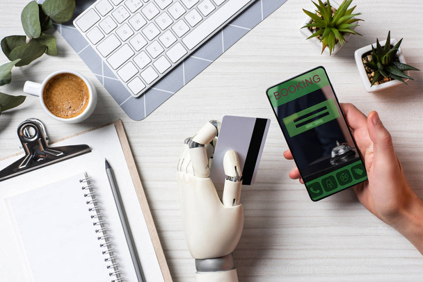 частичный взгляд бизнесмена с протезной рукой, держащего кредитную карту и использующего смартфон с бронированием на экране за столом в офисе
  - Фото, изображение