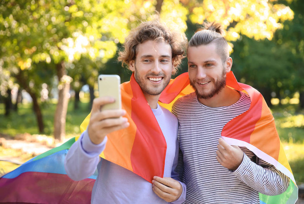 Счастливая гей-пара делает селфи с радужным ЛГБТ-флагом в парке
 - Фото, изображение