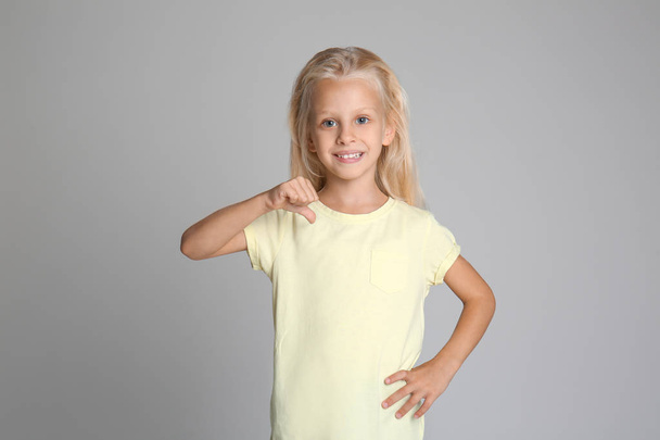 Милая маленькая девочка, указывающая на свою футболку на сером фоне
 - Фото, изображение