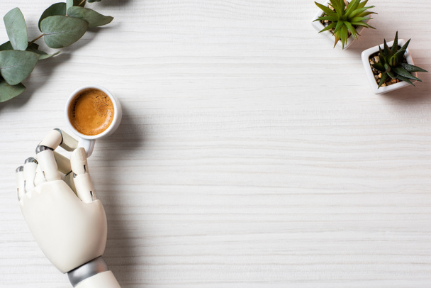visão parcial da mão ciborgue sentada à mesa com xícara de café e plantas no escritório
  - Foto, Imagem