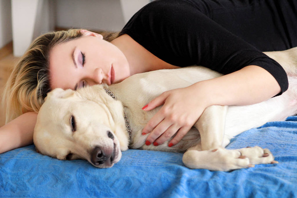Γυναίκα με χαριτωμένα σκυλιά στο σπίτι. Όμορφο κορίτσι ανάπαυσης και ύπνου με το σκύλο στο κρεβάτι στην κρεβατοκάμαρα. Ιδιοκτήτη και σκύλου στον ύπνο στον καναπέ. Κίτρινο Λαμπραντόρ Ριτρίβερ χαλαρώσουν. Το πορτρέτο της γυναίκας και τον καλύτερο φίλο. - Φωτογραφία, εικόνα