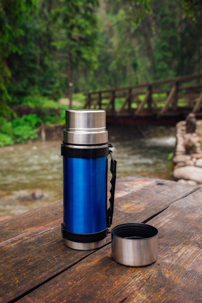 Vista sul thermos blu sul tavolo di legno sul sentiero di trekking. Pausa per bevanda calda
.  - Foto, immagini
