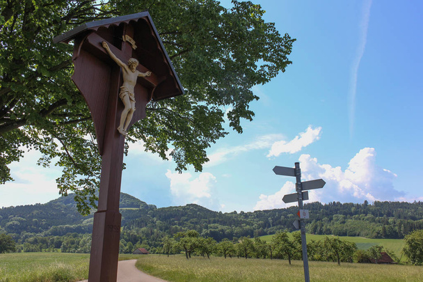 on a very sunny day in june in south germany you see summer landscape with road and crucifix and shields for ways and blue sky on the horizon with cumulus clouds - Photo, Image