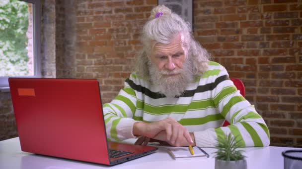 Cute caucasian old man is sitting next to his computer and writing notes, white hair and beard, casual and chill illustration, brick background - 映像、動画