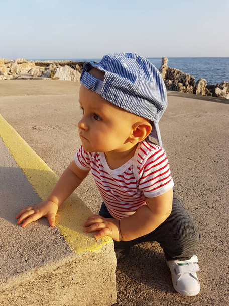 Симпатичный мальчик 1 год носит кепку задом наперёд и полосатую одежду, крупным планом портрет. Средиземное море на заднем плане
 - Фото, изображение