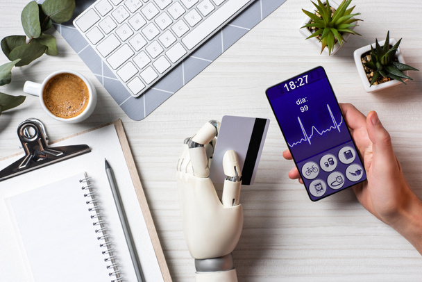 częściowy widok biznesmen cyborg ręką trzyma karty kredytowej i za pomocą smartfona z aplikacją medycznych na ekranie przy stole z filiżanki kawy w biurze  - Zdjęcie, obraz