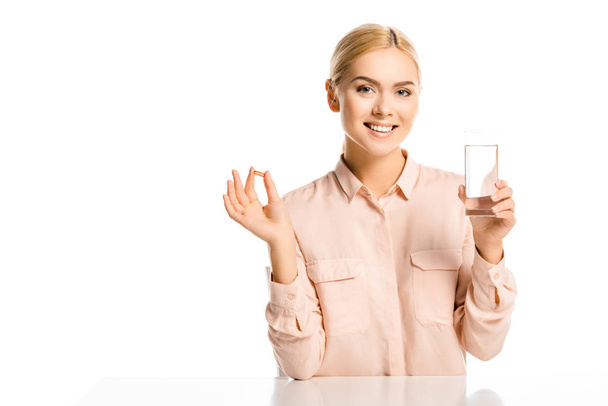усміхнена приваблива жінка тримає склянку води і таблетки ізольовано на білому, дивлячись на камеру
 - Фото, зображення