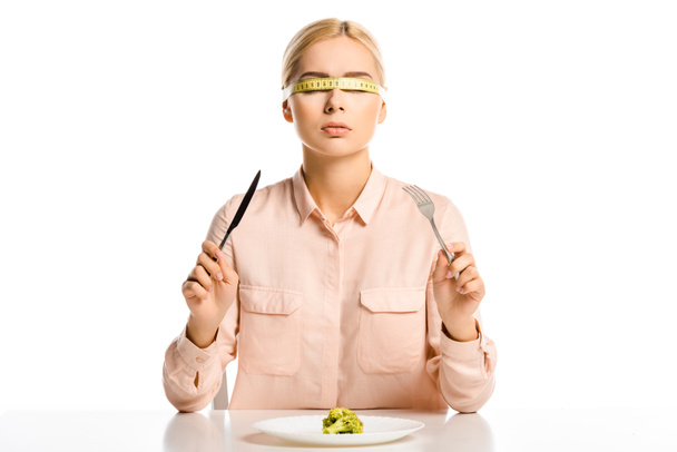 atractiva mujer con cinta métrica en los ojos sosteniendo tenedor y cuchillo, pedazo de brócoli en la placa aislada en blanco
 - Foto, imagen