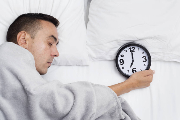 high angle of a young caucasian man in bed, covered with a soft gray blanket, grabbing a clock at seven - Photo, Image