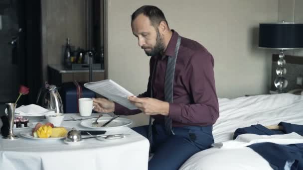 Young businessman reading magazine and drinking tea during breakfast in hotel - Filmati, video