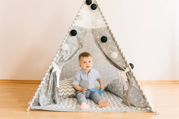 Cute one year old baby playing in a tent, wigwam. Shadow depth of field, natural light. The interior of the children's room is grey, blue and white - Photo, image