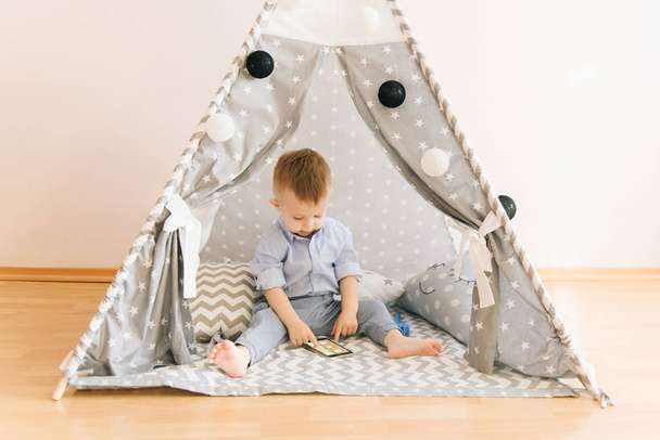 Cute one year old baby playing with smartphones a tent, wigwam. Shadow depth of field, natural light. The interior of the children's room is grey, blue and white - Photo, Image