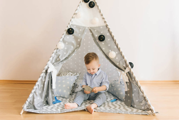 Cute one year old baby playing with smartphones a tent, wigwam. Shadow depth of field, natural light. The interior of the children's room is grey, blue and white - Fotoğraf, Görsel