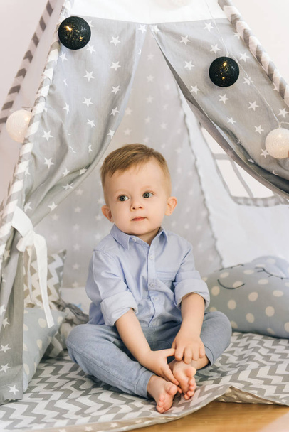 Cute one year old baby playing in a tent, wigwam. Shadow depth of field, natural light. The interior of the children's room is grey, blue and white - Foto, Imagen