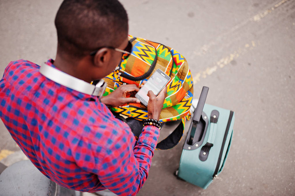African american man in geruite shirt, zonnebril en koptelefoon met koffer en backpack. Zwarte man reiziger mobiele telefoon bij de hand houden. - Foto, afbeelding