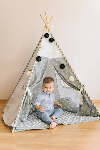 Cute one year old baby playing in a tent, wigwam. Shadow depth of field, natural light. The interior of the children's room is grey, blue and white - Foto, imagen
