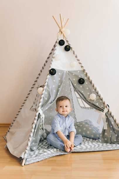 Cute one year old baby playing in a tent, wigwam. Shadow depth of field, natural light. The interior of the children's room is grey, blue and white - Φωτογραφία, εικόνα