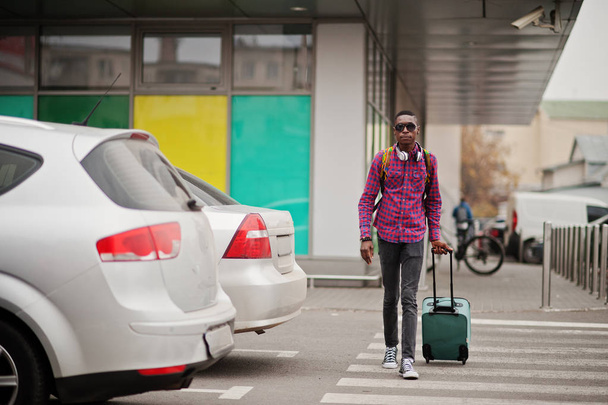African american man in checkered shirt, sunglasses and earphones with suitcase and backpack. Black man traveler walking on crosswalk. - Foto, immagini