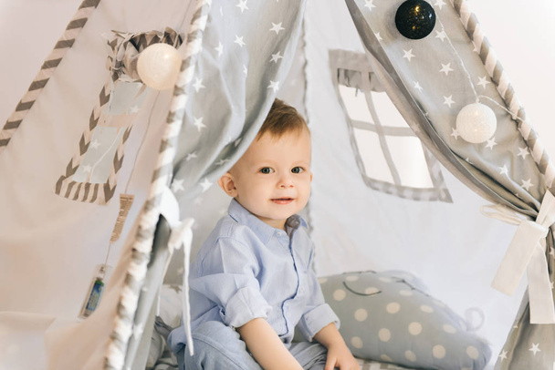 Cute one year old baby playing in a tent, wigwam. Shadow depth of field, natural light. The interior of the children's room is grey, blue and white - Foto, afbeelding