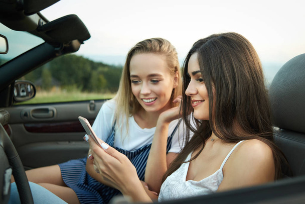 Adorable girls sitting on front seats of cabriolet and looking at phone. - Foto, imagen