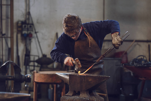 junger Metallbauer oder Schmied, der in einer Werkstatt mit glühend heißem Metall arbeitet und es mit einem Hammer auf dem Amboss zu einer Spule formt - Foto, Bild