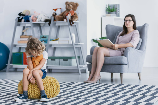 A withdrawn child with an autism spectrum disorder sitting on a pouf during an evaluation session with a professional therapist in an office. - Фото, изображение
