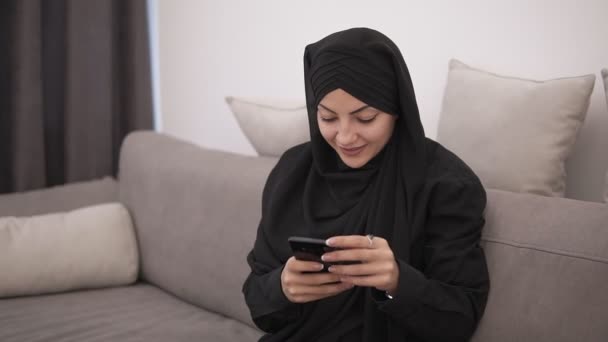 Beautiful muslim woman in black hijab sitting on the sofa and looking to her smartphone and smiling. Modern lofr interior room with white wall on the background - Materiaali, video