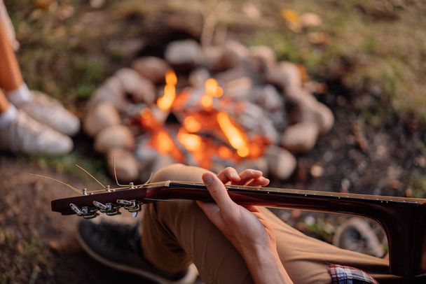 Close up Young man playing guitar near bonfire at sunset on hike - Фото, изображение