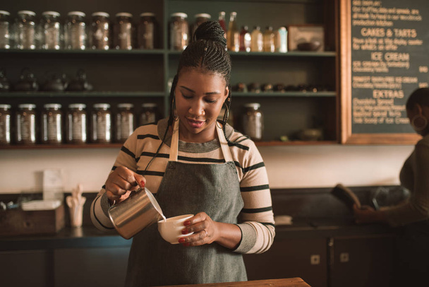 Smiling young African barista pouring frothed milk into a cup of cappuccino behind the counter of her trendy cafe - Fotó, kép
