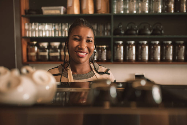 Porträt eines jungen afrikanischen Baristas, der freundlich lächelnd hinter der Bestelltheke eines trendigen Cafés steht  - Foto, Bild