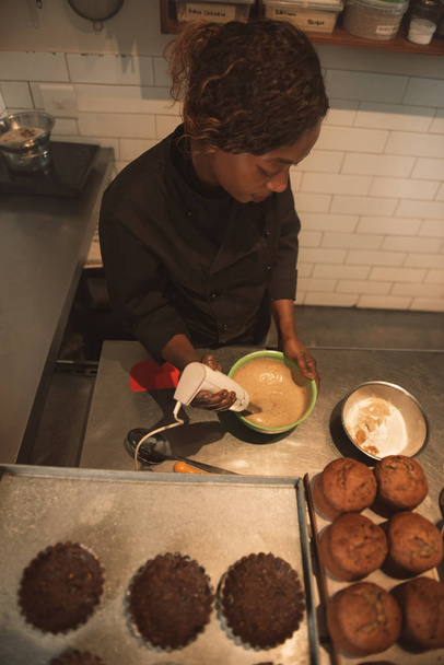Hoge hoek van een Afrikaanse vrouwelijke Bakker mixen van cake beslag terwijl staande bij de balie van een commerciële bakkerij - Foto, afbeelding