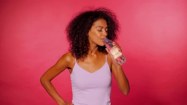 Attractive african-american sporty young woman drinking bottle of water on pink wall background. Girl in sports clothes after fitness. Studio footage. 4k - Video