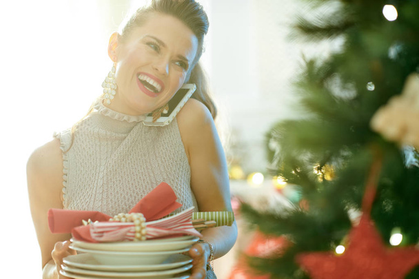 smiling trendy housewife with serving plates talking on smartphone near Christmas tree - Foto, immagini
