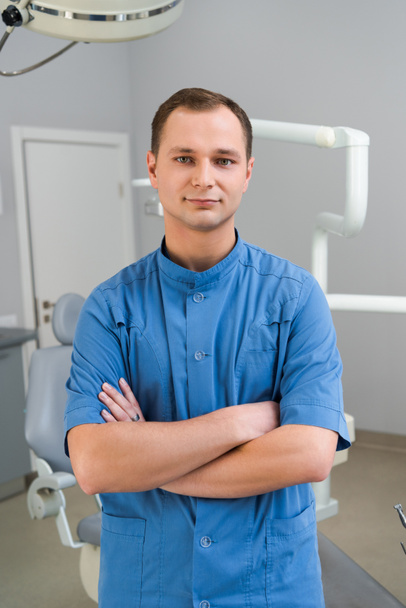 handsome young dentist with crossed arms looking at camera in office - Foto, afbeelding