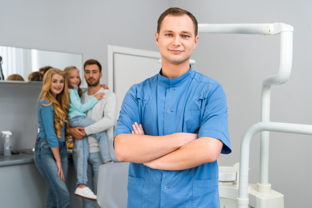 knappe jonge tandarts met gekruiste armen camera terwijl jonge familie staande op achtergrond samen kijken - Foto, afbeelding